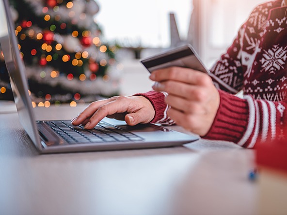 person wearing Christmas jumper using credit card with laptop, with decorated Christmas tree in background